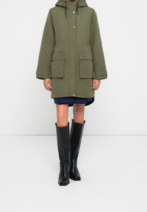 Femme portant un parka à capuche vert olive avec de grandes poches, une jupe bleue et des bottes en cuir noires montantes jusqu'aux genoux, debout sur un fond uni.