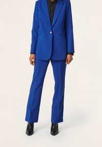 Modrý blazer a odpovídající kalhoty z texturované tkaniny. Zapínání blazera na knoflíky, v kombinaci s černou košilí s límečkem a černými kotníkovými botami.