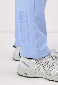 Light blue sweatpants made of cotton blend fabric, featuring a side print. Paired with grey athletic shoes, showcasing a silver logo and textured sole.