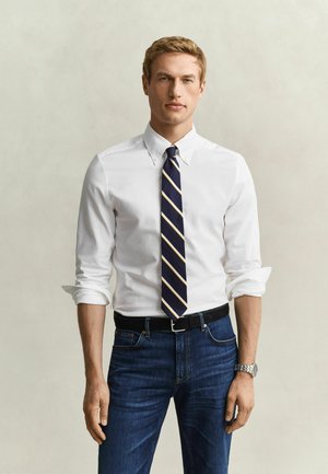 Jeune homme aux manches retroussées, portant une chemise blanche à boutons, une cravate rayée bleu marine, un jean bleu, une ceinture noire et une montre-bracelet argentée.
