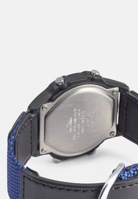 Relógio digital com fundo em aço inoxidável, caixa de plástico preto, correia trançada azul e dois botões de cada lado. Apresenta uma fivela prateada.