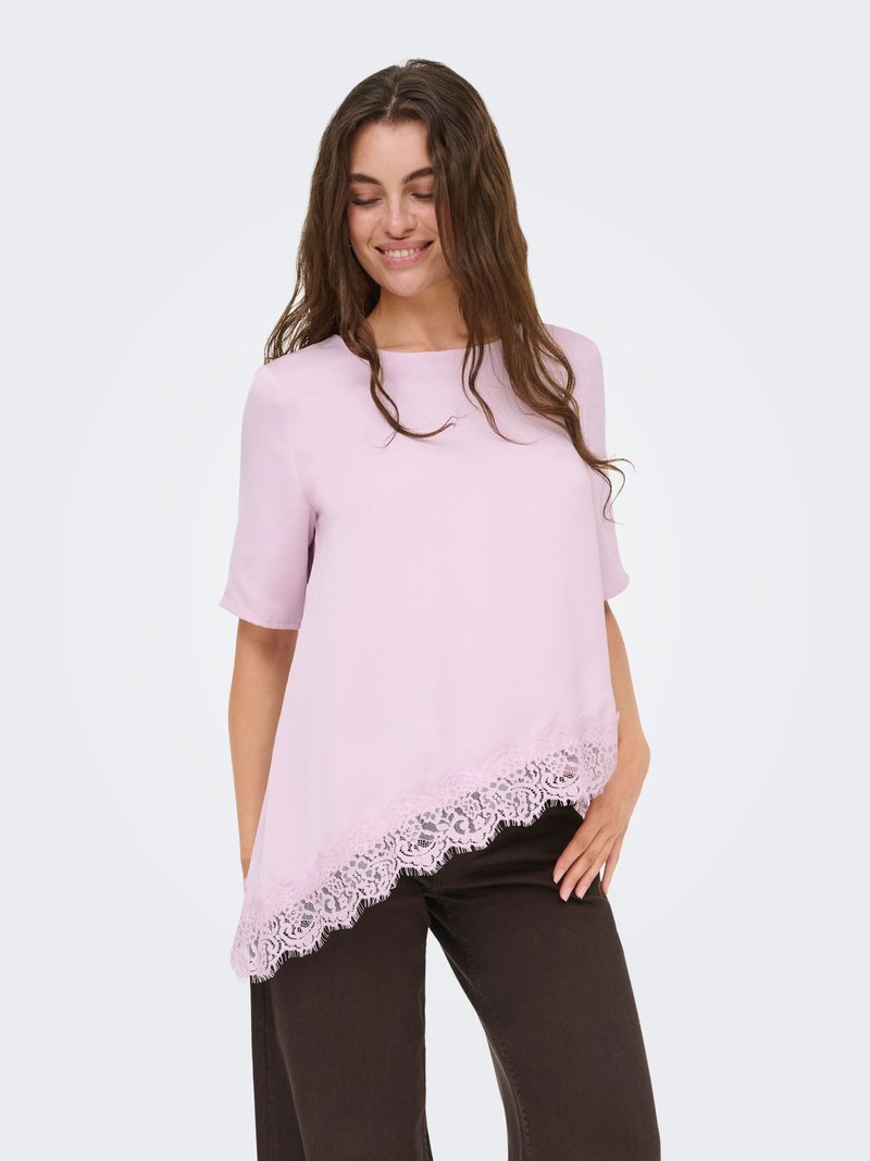 Femme aux longs cheveux bruns portant un haut asymétrique rose clair avec bordure en dentelle et pantalon marron foncé, souriante les yeux fermés.