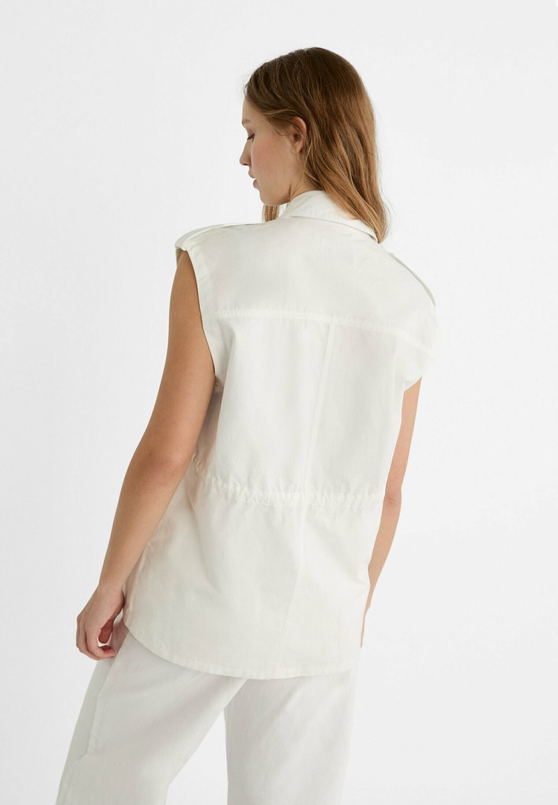 Chemise sans manches beige clair avec un col pointu, des coutures ton sur ton et un détail de taille froncée, vue de dos sur fond blanc.