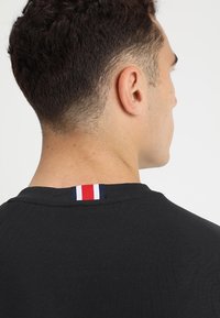 Camiseta negra con escote redondo, que presenta una etiqueta tricolor en la parte posterior, con franjas rojas, blancas y azules.