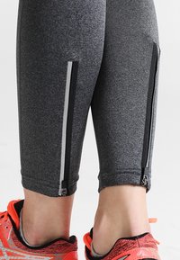 Šedé sportovní legíny s zipy na kotnících a reflexními proužky, vyrobené z strukturovaného, elastického materiálu, spojené s jasnými běžeckými botami.