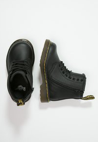 Botine negre din piele cu cusături galbene, vârf rotund, șireturi în față și fermoar pe lateral. Au o talpă din cauciuc texturată și un eticheta a mărcii.