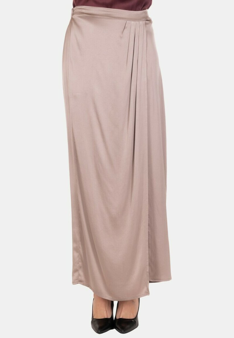 Gonna lunga beige da donna con pieghe sottili su un lato, indossata con tacchi neri a punta, su uno sfondo semplice.