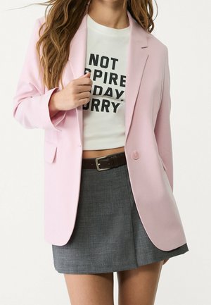 Kobieta ubrana w jasnoróżową marynarkę na białym krótko przyciętym T-shircie z grafiką oraz szarej mini spódnicy z paskiem, pozująca na jednolitym białym tle.