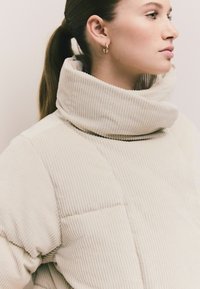 Beige Kordjacke mit hohem Kragen und gestepptem Design. Glatte Textur mit vertikalen Rippungen; bietet eine lockere Passform.