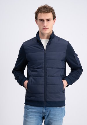 Junger Mann trägt eine marineblaue gesteppte Jacke mit Reißverschluss über einem hellen Pullover, die Hände in den Taschen, und steht vor einem einfarbigen hellen Hintergrund.