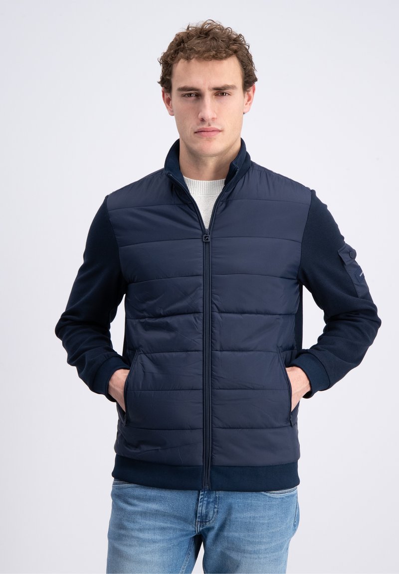 Young man wearing a navy quilted zip-up jacket over a light sweater with hands in pockets, standing against a plain light background.