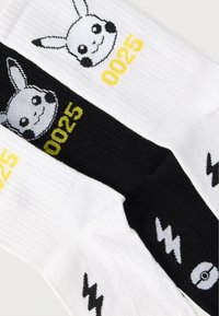 Βαμβακερές κάλτσες με σχέδια Πικάτσου. Δύο χρώματα: λευκό με κίτρινο και μαύρο με γκρι λεπτομέρειες. Περιλαμβάνει αριθμό "0025" και ηλεκτρικά μοτίβα.