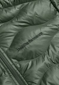 Chaqueta acolchada verde con textura brillante, patrones de costura en forma de ola y logo "Peak Performance" en bordado negro. Cierre de cremallera.