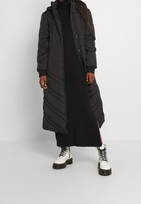 Langer schwarzer Steppmantel mit Reißverschluss vorn, Kapuze und gerippten Bündchen; hat ein diagonales Steppmuster. Wird über einem schwarzen Kleid und weißen Stiefeln getragen.