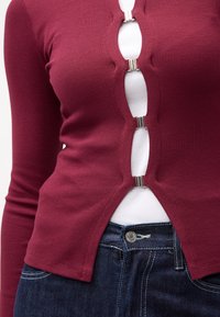 Un top burgundy cu mâneci lungi, caracterizat printr-o textură ribbed, cu deschideri frontale securizate cu elemente metalice argintii, purtat peste o cămașă albă și asortat cu blugi negri.