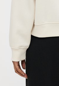 Cremefarbener Sweatshirt mit gerippten Bündchen, zeigt weiche Textur und cropped Design, kombiniert mit einem schwarzen, gerippten Rock.