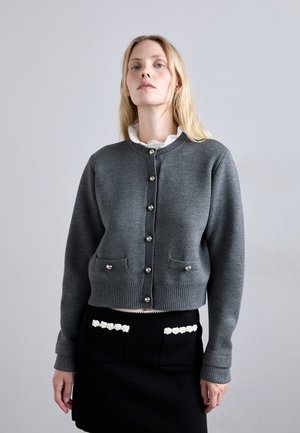 Gray knitted cardigan with a round neckline, button front, and two front pockets. Paired with a black skirt featuring white floral accents.