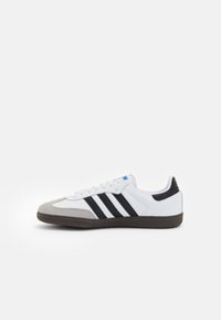 Zapatillas blancas con rayas negras, puntera de ante gris y suela marrón oscuro sobre fondo blanco liso, vista lateral.