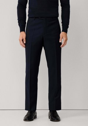 Man wearing black dress pants, black sweater, and black dress shoes standing against a plain light background.