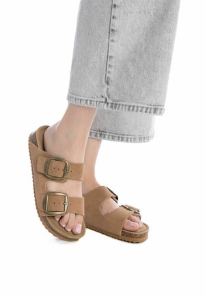 Sandalias de ante marrón con dos correas anchas, que presentan hebillas metálicas grandes, sobre una suela texturizada; combinadas con jeans de pierna ancha gris claro.