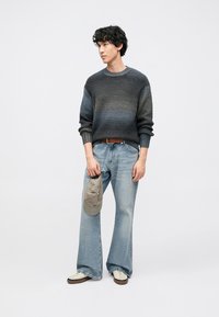 Siv sivani pulover z vzorcem, svetlo modre kavbojke z širokimi hlačnicami, bele superge in bež kapa, držana v roki. Preprost dizajn, priložnostna prileganje.