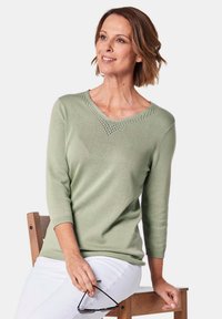 Hellgrüner V-Ausschnitt-Pullover mit 3/4-Ärmeln, ausgestattet mit dekorativen Ausschnittdetails entlang des Halsausschnitts und einer weichen Stricktextur.