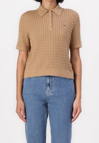 Kobieta w beżowej, teksturowanej, krótkiej koszulce polo, połączonej z dżinsami w wysokim stanie, stojąca na jednolitym tle.