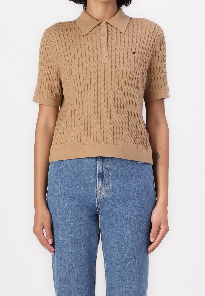 Kobieta w beżowej, teksturowanej, krótkiej koszulce polo, połączonej z dżinsami w wysokim stanie, stojąca na jednolitym tle.