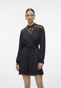 Rochie neagră cu mâneci lungi, având un yoke din dantelă, un cordon legat la talie și o fustă evazată. Fabricată dintr-un material fin și ușor.