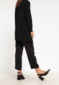 Svart långärmad topp, rak skärning med sidoknytt; kombinerad med svarta, löst sittande byxor och blanka svarta klackskor med blockklack.