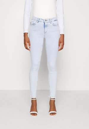 Personne portant un jean skinny bleu clair avec des ourlets effilochés et des talons blancs à bout ouvert, associé à un haut blanc à manches longues.