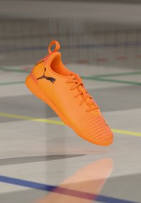 Félfényes narancssárga Puma teremfoci cipő, ami felfüggesztve lebeg egy színes vonalakkal jelölt tornaterem padlóján.