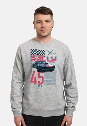 Mężczyzna w szarej bluzie z czarnym samochodem wyścigowym, szachownicą, czerwonym napisem "Gotham Rally" oraz dużą czerwoną liczbą 45 z przodu.