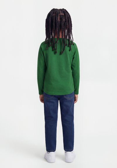 Bambino con trecce lunghe che indossa una maglia verde a maniche lunghe, jeans blu e sneakers bianche, in piedi di spalle su uno sfondo bianco.