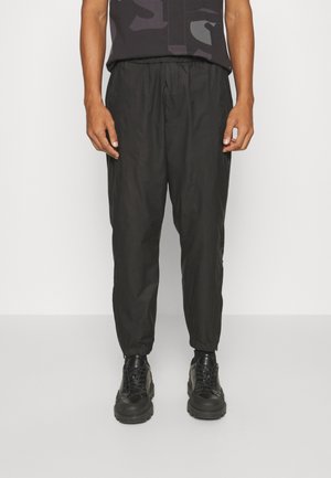 Mann trägt schwarze Jogginghosen mit elastischem Bund und elastischen Bündchen sowie schwarze klobige Schnürschuhe, steht vor einem einfarbigen Hintergrund.