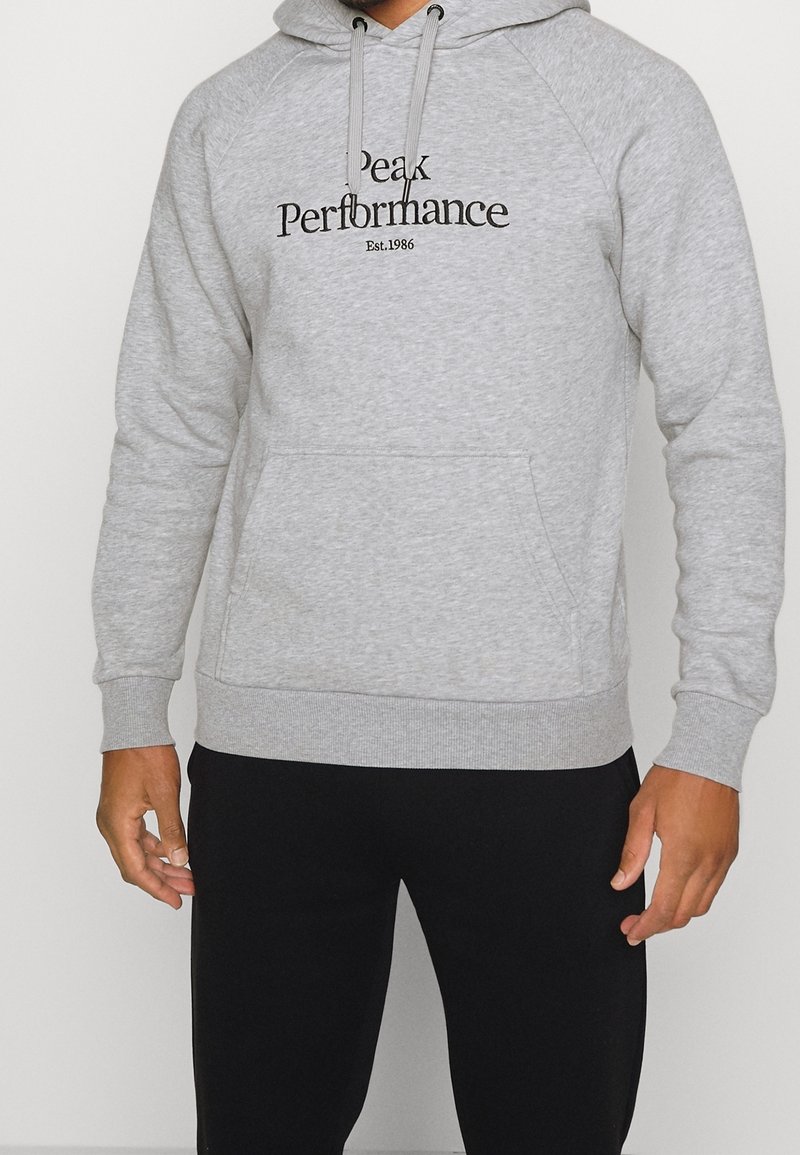 Homme portant un sweat à capuche gris clair avec le texte "Peak Performance Est. 1986" et un pantalon noir, debout les bras détendus le long du corps.
