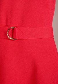 Punainen kangasmekko, jossa on teksturoitu pinta, yhteensopiva vyö ja kultainen yksityiskohta soljessa. Malli on vartalonmyötäinen vyötäröltä.