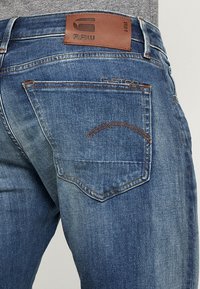 Jeans i mellanmörk blå denim med en blekt utseende. Har en brun läderlogotyp på midjebandet och böjd sömnad på bakfickorna.