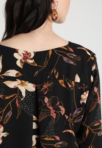 Woman with dark hair wears black floral blouse with orange, pink, and white flowers and tortoiseshell hoop earring with gold charm.