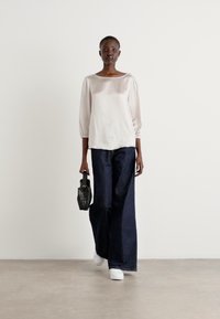 Light pink blouse with three-quarter sleeves, paired with wide-leg dark denim jeans. Model holds a small black textured handbag and wears white shoes.