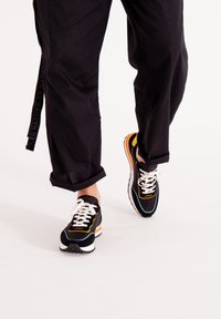 Sneakers atletici neri con lacci bianchi, dettagli arancioni e gialli, e una suola texturizzata. Indossati con pantaloni neri larghi.
