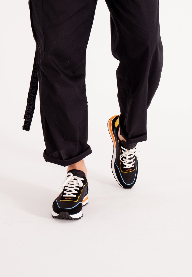 Sneakers atletici neri con lacci bianchi, dettagli arancioni e gialli, e una suola texturizzata. Indossati con pantaloni neri larghi.