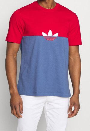 Mand iført en kortærmet t-shirt med en rød øverste halvdel og en blå nederste halvdel, med et hvidt og rødt logo på brystet, kombineret med hvide bukser.