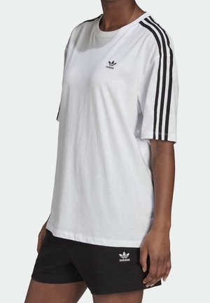 Osoba w białej koszulce Adidas z czarnymi trzema paskami na rękawach i czarnych szortach Adidas.