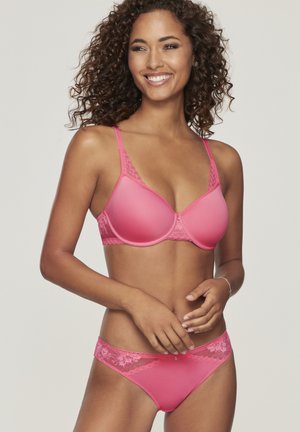 Frau mit lockigem Haar, die lächelt, trägt ein rosa, mit Spitze verziertes BH und passende Unterwäsche vor einem schlichten hellen Hintergrund.