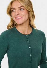 Žena s blond vlasmi, oblečená v zelenej gombíkovej kardigánke, sa usmieva a pozerá na stranu na pozadí jednofarebnej bielej plochy.