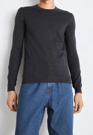 Mand iført en tætsiddende mørkegrå langærmet sweater og løse blå cowboybukser står mod en almindelig lys væg.