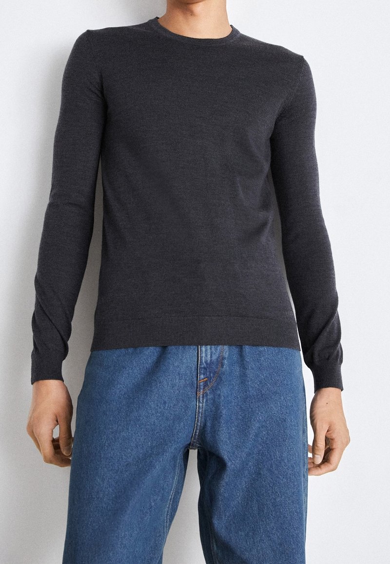 Homme portant un pull ajusté gris foncé à manches longues et un jean bleu ample, debout contre un mur clair uni.