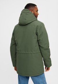 Hombre con una chaqueta de invierno verde con capucha y jeans azules, visto desde atrás contra un fondo blanco liso.