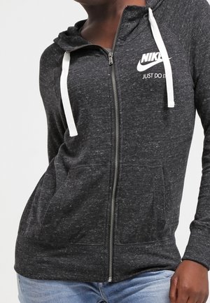 Ciemnoszara bluza Nike z kapturem i zamkiem błyskawicznym, z białymi sznurkami i napisem „JUST DO IT”, noszona z jasnoniebieskimi dżinsami.